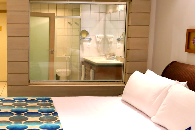 imagem do quarto com uma cama de casal e o banheiro com uma parede de vidro