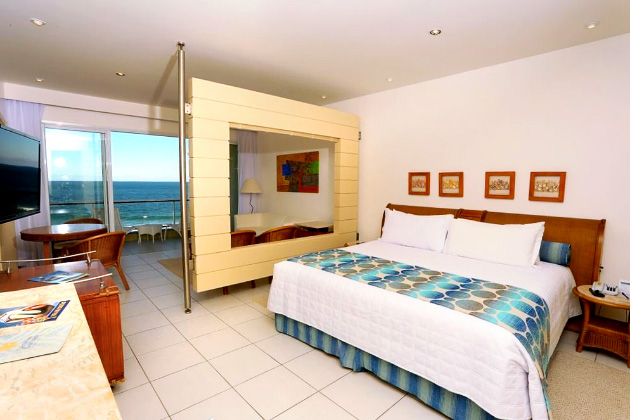 Imagem do quarto com uma cama de casal e uma tv, com vista para o mar