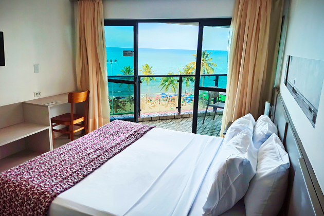 Imagem do quarto com uma sacada com uma vista para a praia contendo uma cama de casal e uma tv
