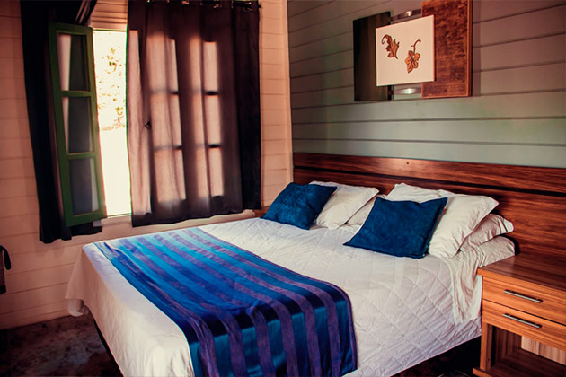 Imagem de uma cama de casal, uma janela com cortina e moveis ao lado da cama.