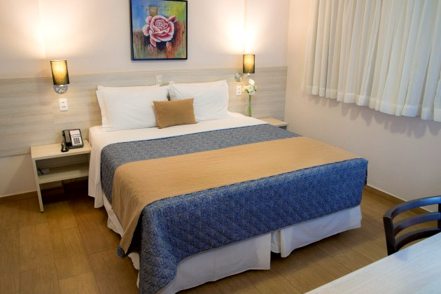 Imagem de uma cama de casal com lençol azul e duas luminárias em cada lado