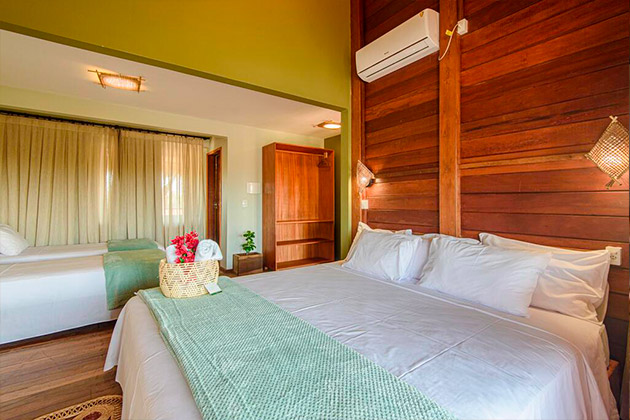 imagem de uma cama de casal com a parede de fundo em madeira, ar condicionado e mais duas camas de solteiro.